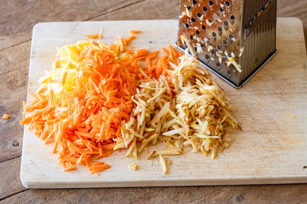 Grated carrots and grated apple on a wooden board next to a grater.