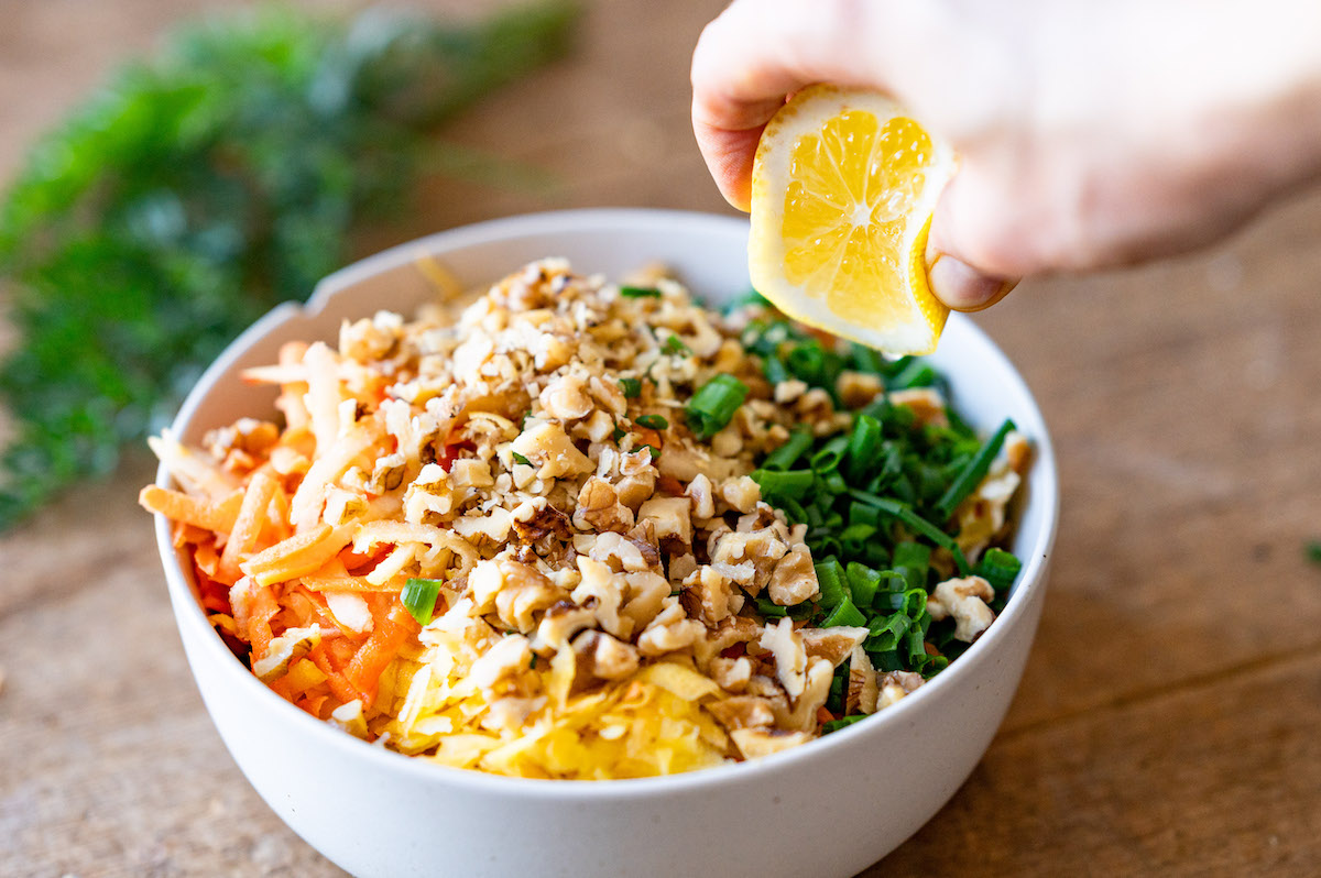 A lemon is being squeezed so that the juice is being poured over all carrot salad ingredients.