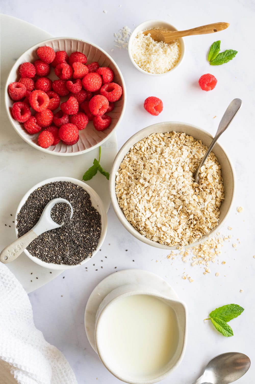 Overhead shot of ingredients for the overnight oats: Rolled oats, chia seeds, coconut flakes, milk and raspberries.