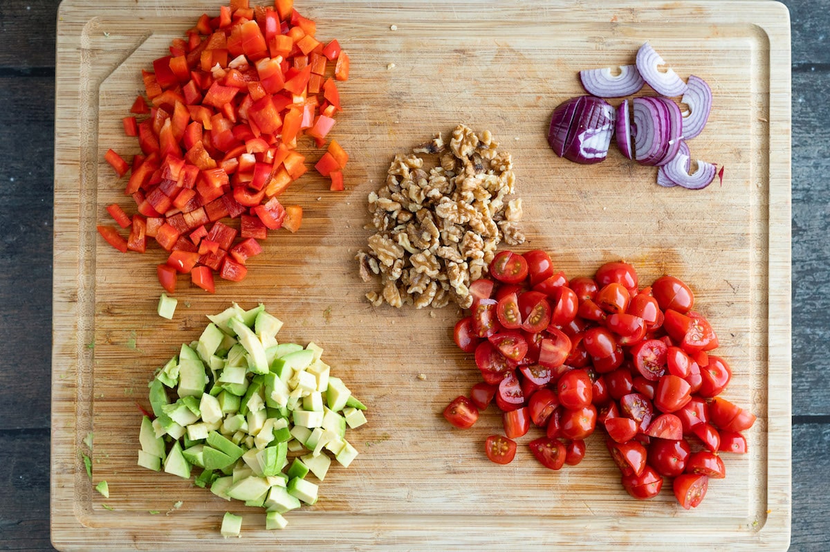 Ingredients for the red lentil pasta salad chopped on a board: bell pepper, walnuts, avocado, red onions and tomatoes.