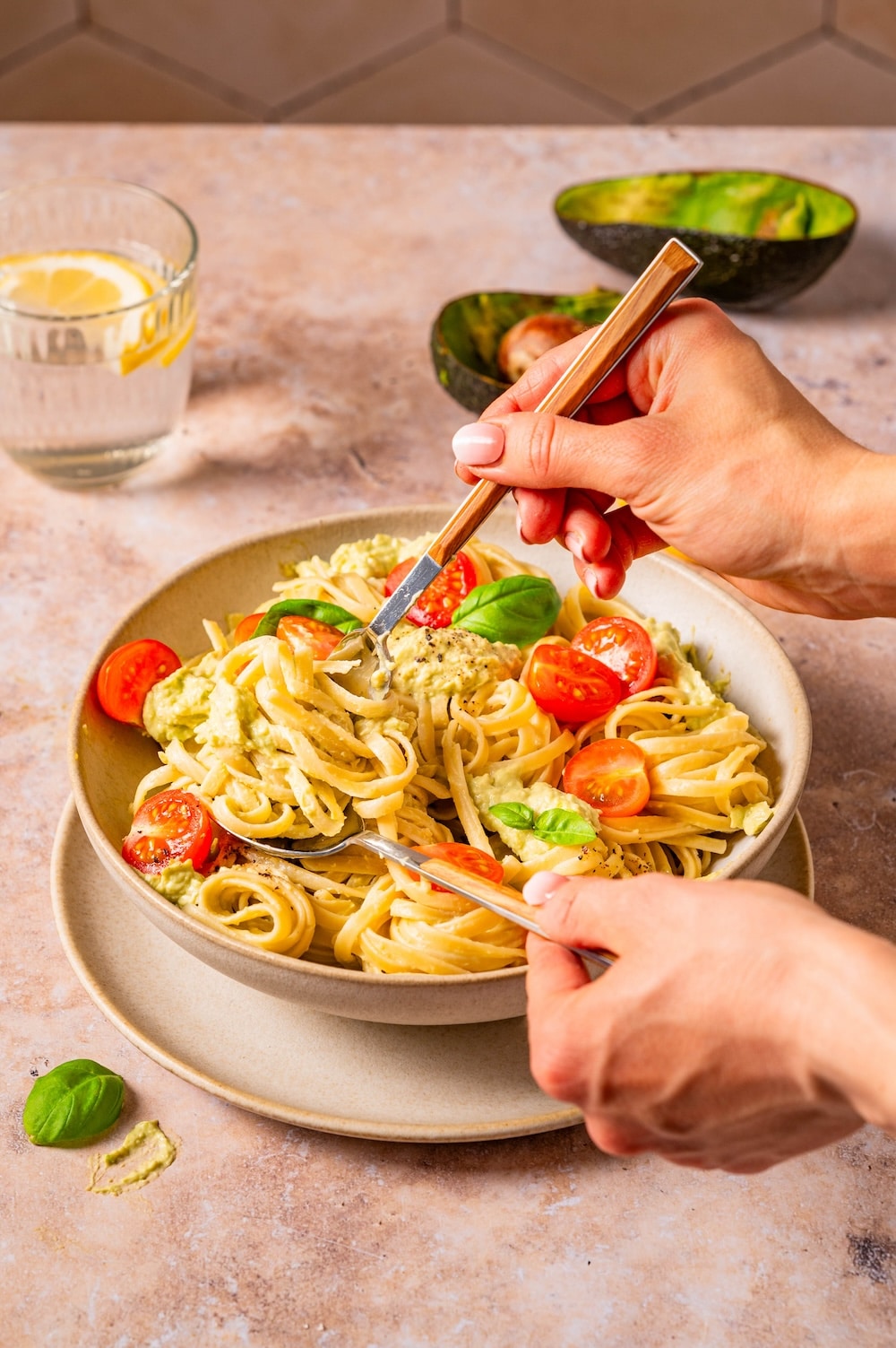 Avocado and tomato pasta in a bowl, being forked up with fork and spoon.