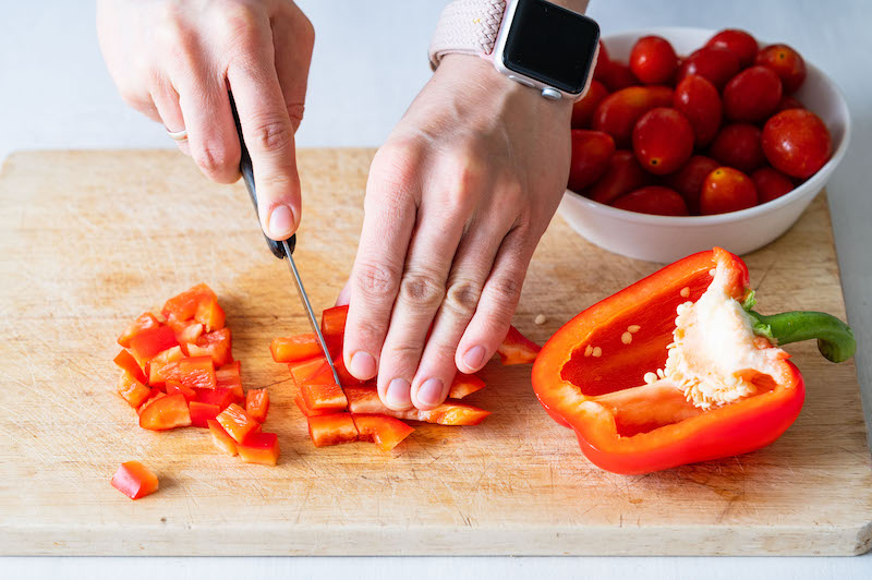 A red bell pepper is being cut into small chunks. Cherry tomatoes in the background.
