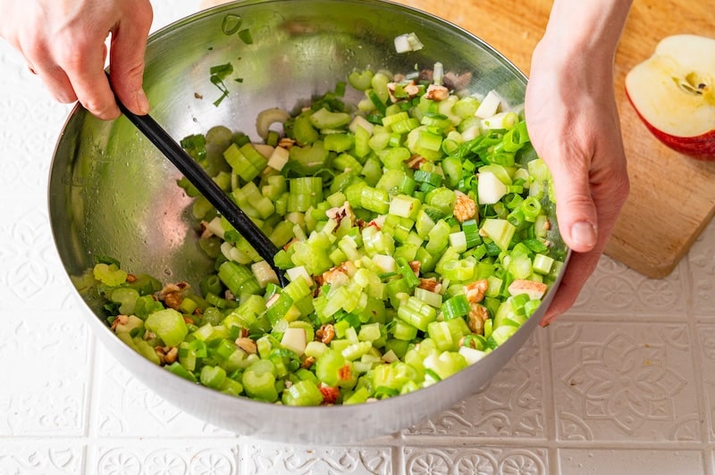 Chopped celery, apple, scallions and walnuts are being mixed in a bowl.