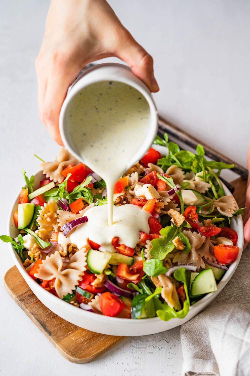 Yogurt dressing is being poured over a wholegrain pasta salad in a bowl.
