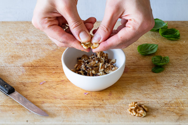 Walnuts are being crumbled by hand. There's a few basil leaves in the background.